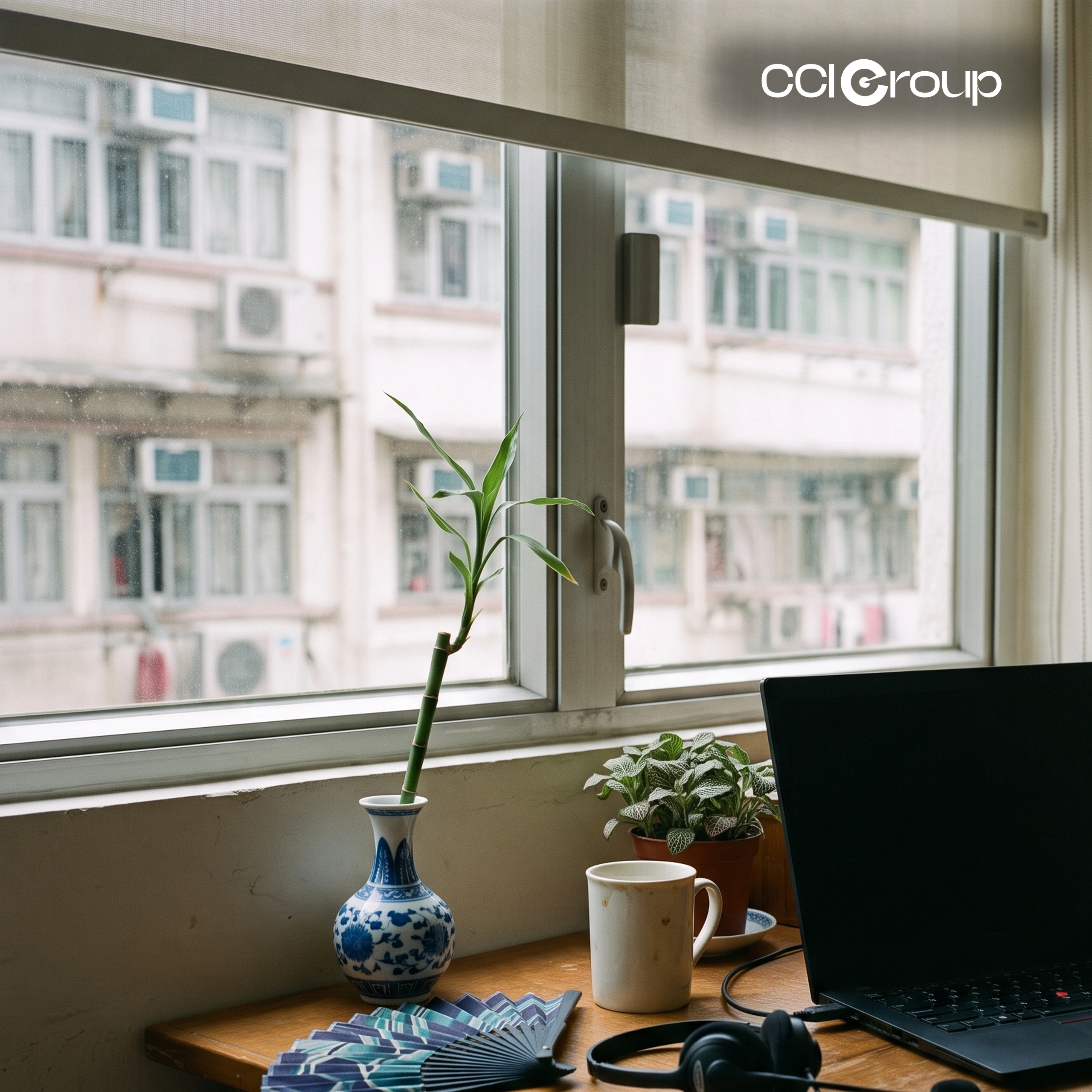 Wooden desk with laptop, black headphones, blue-and-white porcelain vase with lucky bamboo, and folding fan by a city window.