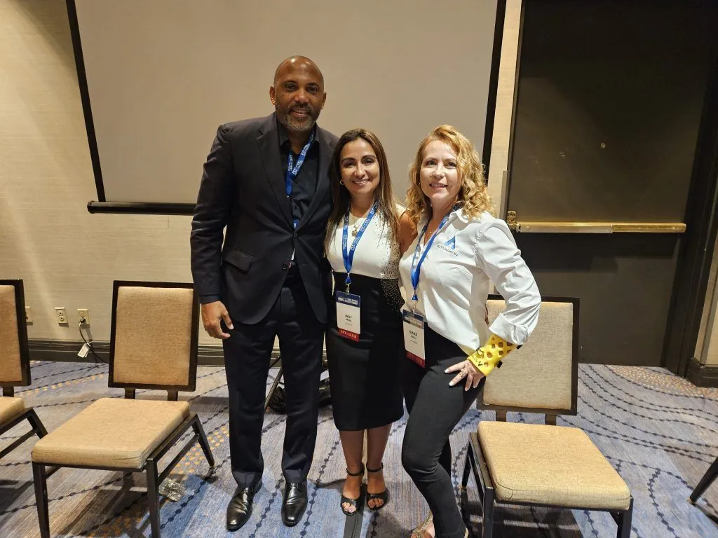 Three attendees wearing lanyards and name badges posing together in a conference room with beige chairs