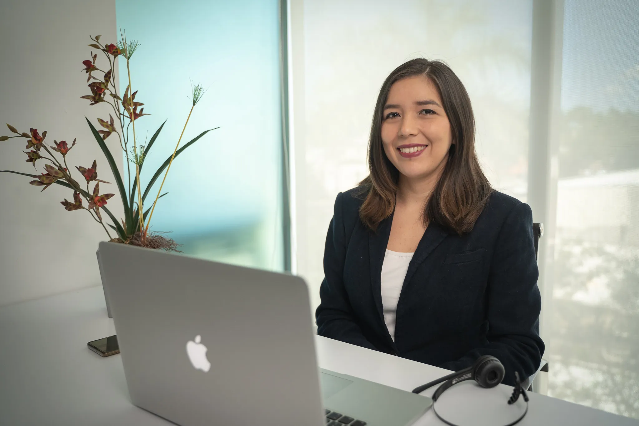 Smiling woman in a dark blazer seated at a white desk with a laptop, headset and plant