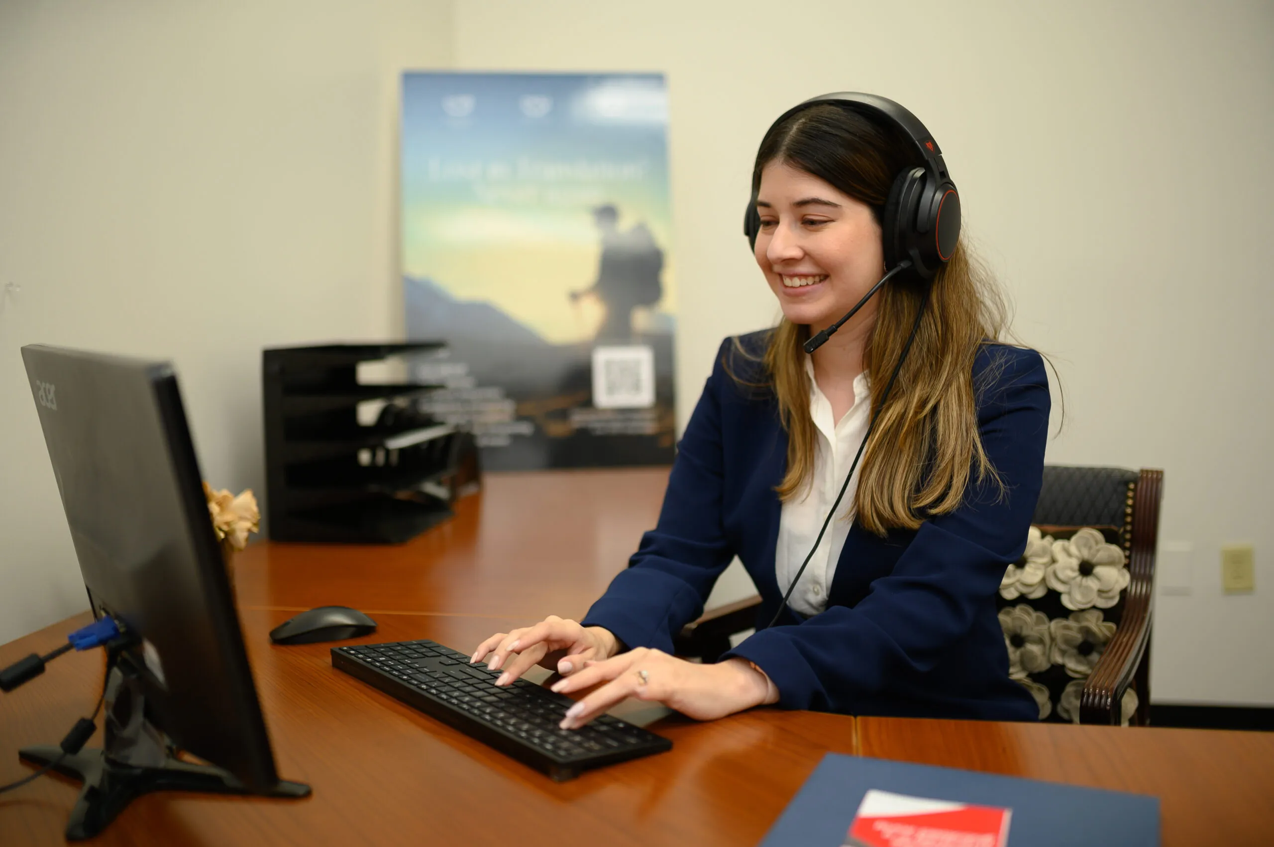 Smiling woman in a navy blazer wearing a headset with microphone while typing at a desktop computer